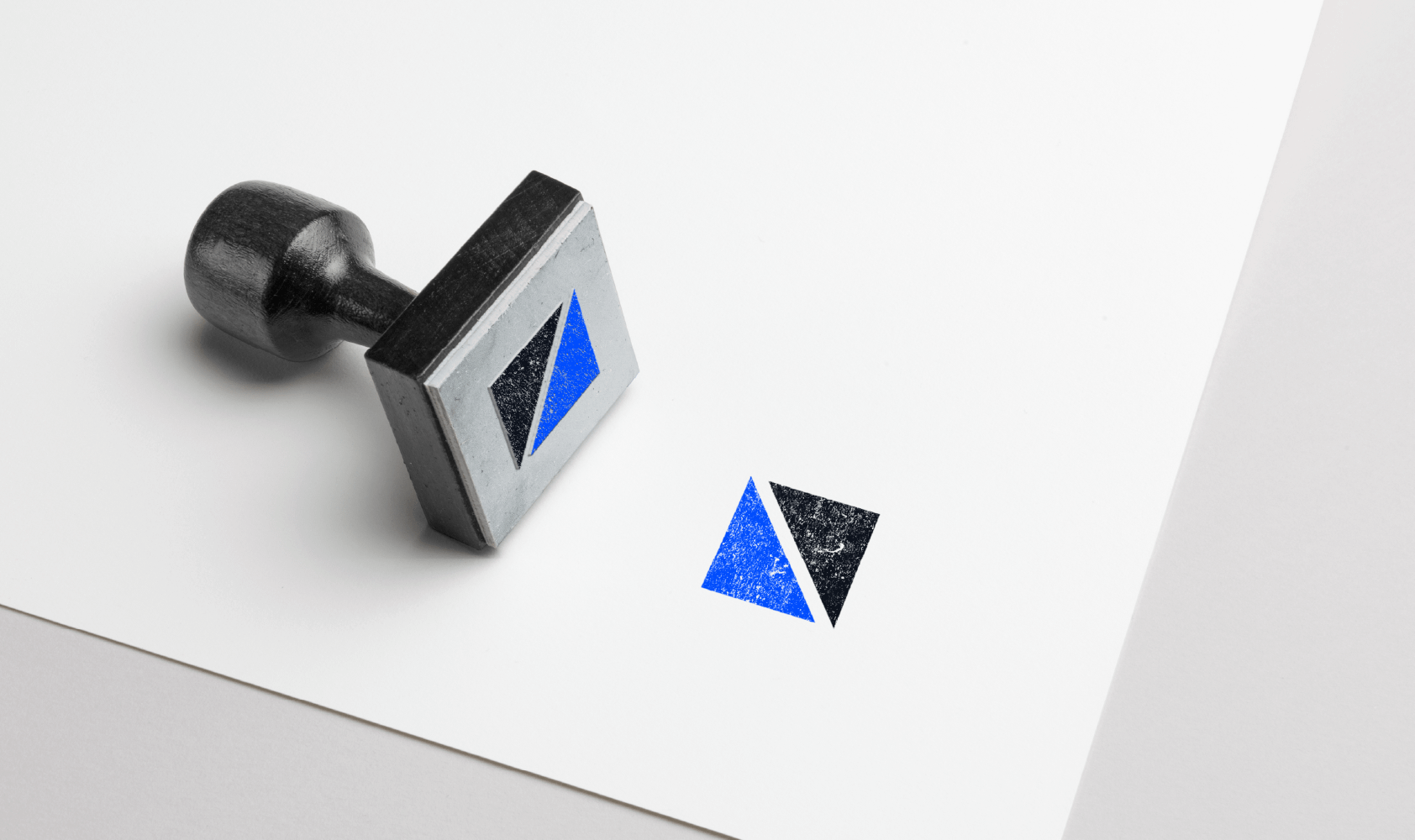 A rubber stamp with a split square design—blue triangle on the left, black triangle on the right—rests on white paper, leaving a matching stamped impression beside it.